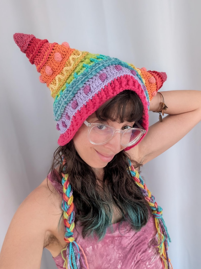 May include: A colorful, handmade crochet hat in a rainbow of hues, featuring a pointed top and textured details. The hat has a pink brim and is adorned with small, round accents. Braided hair extensions in matching colors complete the look.