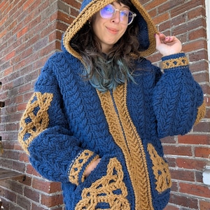 May include: A handmade blue hooded jacket with a gold-colored trim and accents. The jacket features a cable knit design, a zipper closure, and two front pockets. The hood and cuffs are trimmed with a contrasting gold color.