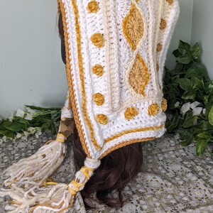 May include: White crochet hooded scarf with a yellow and white pattern and tassels. The scarf is made with a chunky yarn and has a textured design.