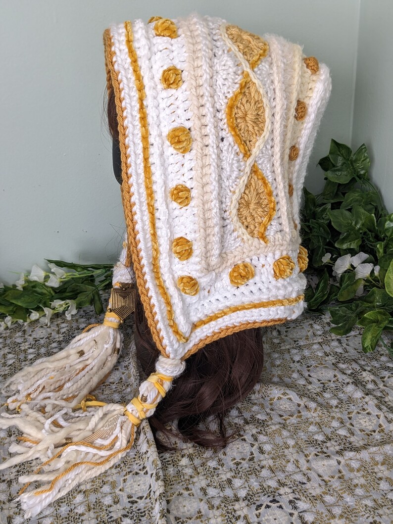 May include: White crochet hooded scarf with a yellow and white pattern and tassels. The scarf is made with a chunky yarn and has a textured design.