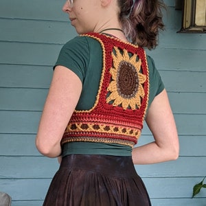 Puede incluir: Un chaleco de ganchillo con forma de girasol hecho con hilo rojo, naranja, amarillo y marr&oacute;n. El chaleco tiene forma cuadrada con un gran dise&ntilde;o de girasol en el centro.