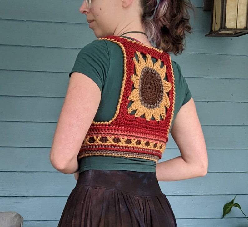 Puede incluir: Un chaleco de ganchillo con forma de girasol hecho con hilo rojo, naranja, amarillo y marr&oacute;n. El chaleco tiene forma cuadrada con un gran dise&ntilde;o de girasol en el centro.