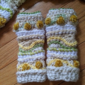 May include: A pair of crocheted arm warmers with a white base and yellow, green, and brown accents. The arm warmers have a textured design and are decorated with small velvet pom-poms.