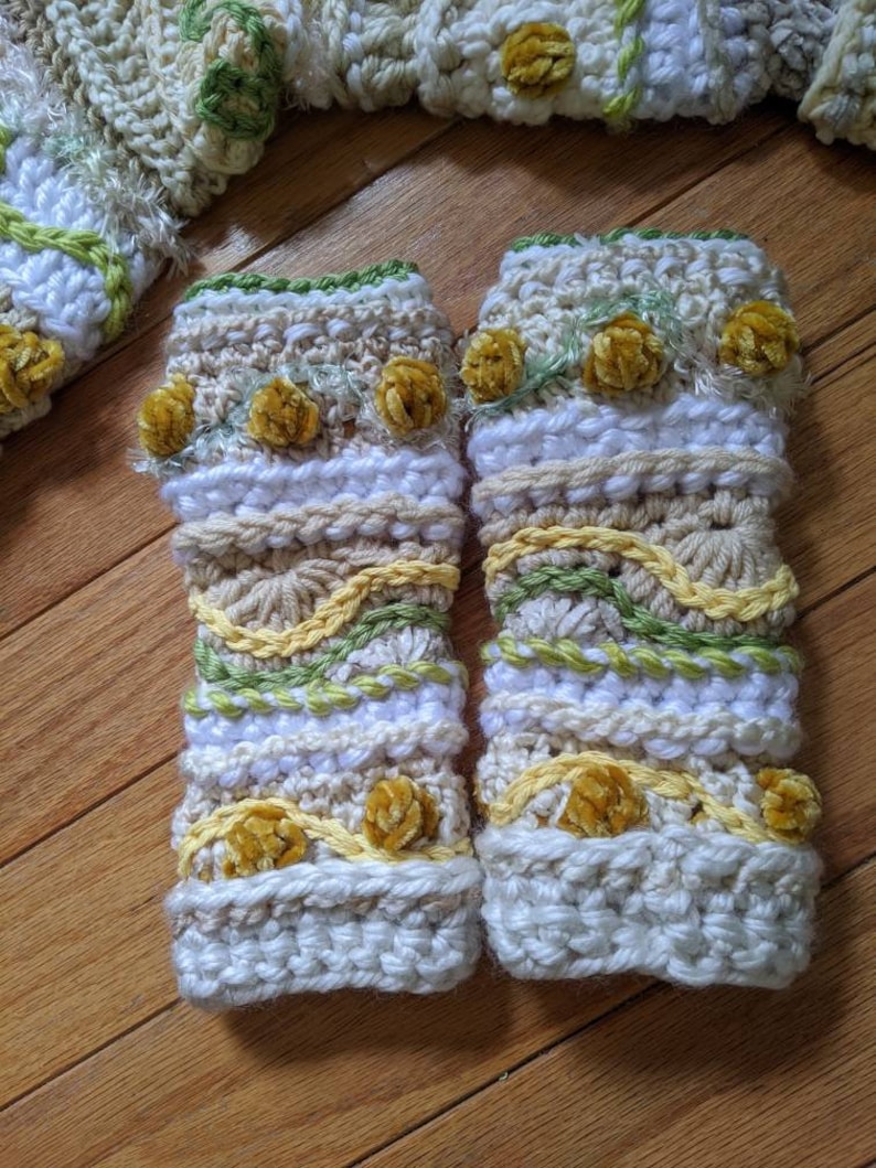 May include: A pair of crocheted arm warmers with a white base and yellow, green, and brown accents. The arm warmers have a textured design and are decorated with small velvet pom-poms.