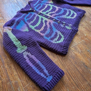 May include: A purple crochet cardigan with a skeleton design. The skeleton is made with pastel rainbow colors. The cardigan has long sleeves and a button-up front. The buttons are silver. The cardigan is made of a thick yarn.
