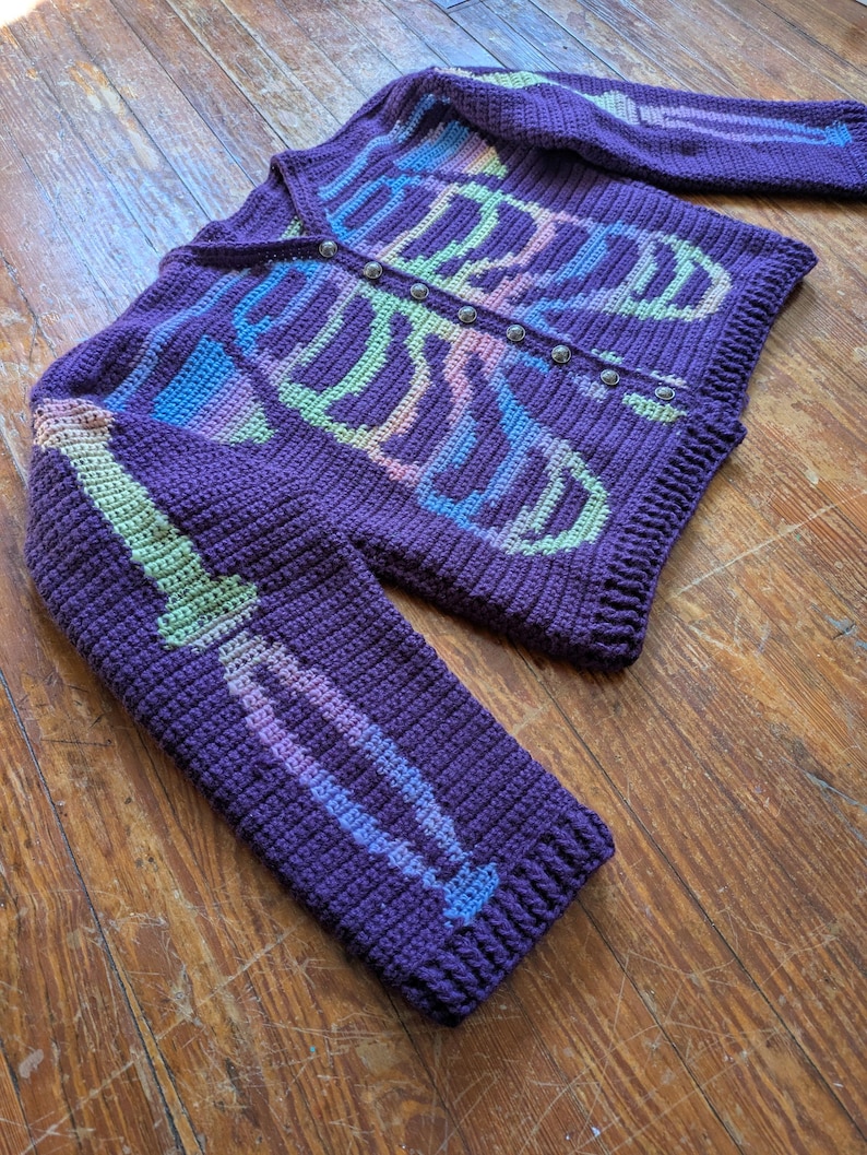 May include: A purple crochet cardigan with a skeleton design. The skeleton is made with pastel rainbow colors. The cardigan has long sleeves and a button-up front. The buttons are silver. The cardigan is made of a thick yarn.