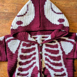 May include: A burgundy and cream crochet skeleton hoodie with a button-up front. The hood and front of the sweater feature a cream-colored skeleton design. The sweater is made of a soft, textured yarn.
