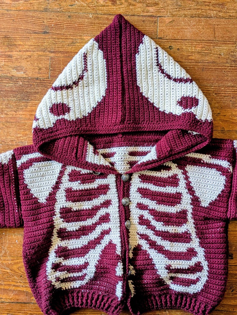 May include: A burgundy and cream crochet skeleton hoodie with a button-up front. The hood and front of the sweater feature a cream-colored skeleton design. The sweater is made of a soft, textured yarn.