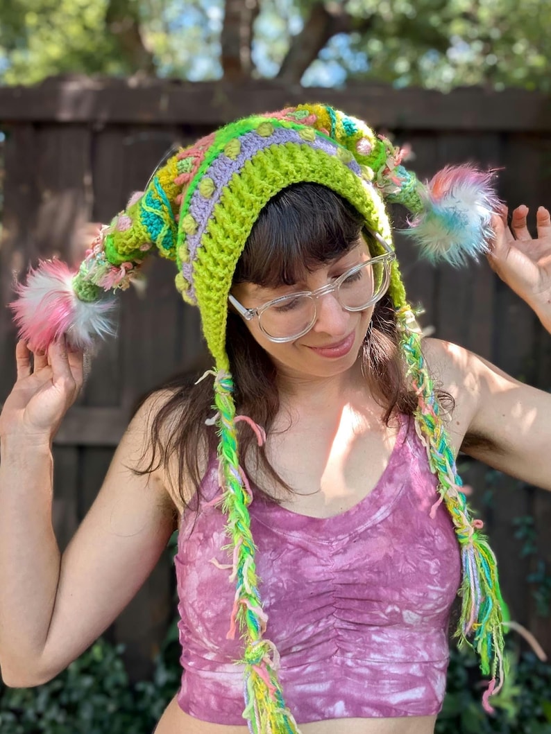 May include: A whimsical, handmade crochet hat in vibrant colors. The hat features a long, pointed shape with a lime green base, accented with lavender, pink, and yellow details. It has dangling braids and fluffy pom-poms at the ends, creating a playful and unique accessory.