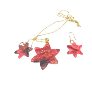 May include: A gold chain necklace and earrings set with red and black star-shaped pendants. The stars have a marbled effect with gold flecks.