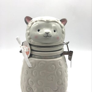 May include: A white ceramic jar shaped like a sheep with a hinged lid. The jar has a cute cartoon face and a swirl pattern on the body.