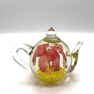 May include: A clear glass teapot with a red and yellow floral design inside. The teapot has a rounded body, a curved handle, and a spout. The design includes red petals and a yellow base.