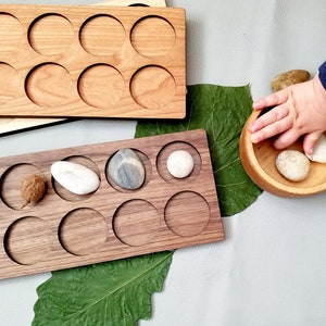 May include: Two wooden trays with circular cutouts, one light wood and one dark wood. The light wood tray has 12 cutouts and the dark wood tray has 10 cutouts. The dark wood tray has a pine cone, a small brown object, and a white rock in the cutouts. A small wooden bowl with three white rocks is on a white surface.