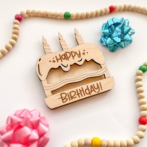 以下が含まれることがあります： ケーキの形をした木製バースデーカードで、「Happy Birthday!」の文字が書かれています。カードは、カラフルなアクセントが付いた木製のビーズの紐で囲まれています。