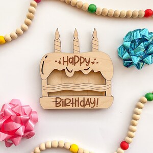 以下が含まれることがあります： ハッピーバースデー！と書かれた3本のろうそくがついた木製の誕生日ケーキの看板。看板はカラフルなビーズが置かれた白い背景の上に置かれています。