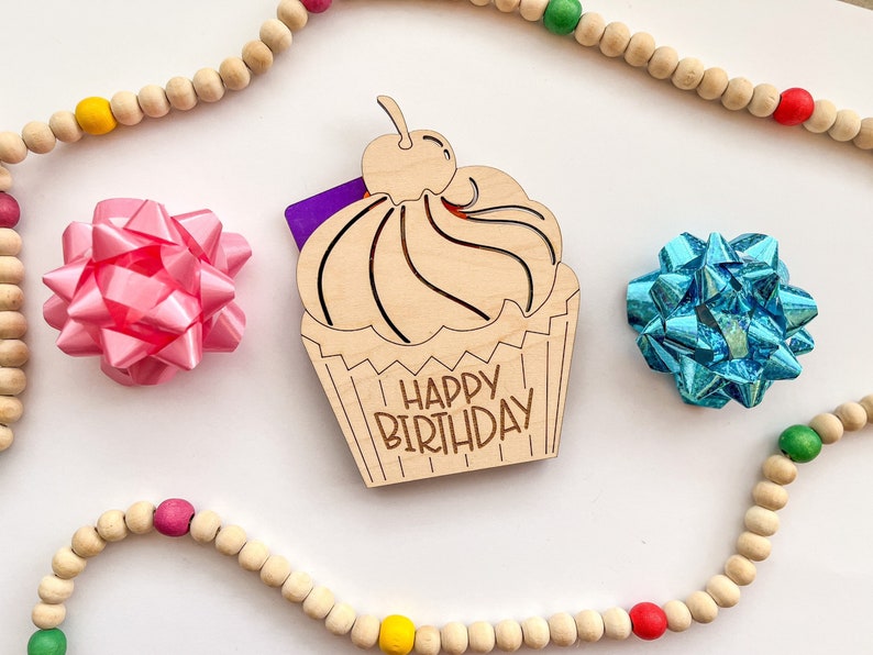 May include: Wooden cupcake card with the words "Happy Birthday" on it. The cupcake is decorated with a cherry and frosting. The card is surrounded by a string of wooden beads with red, yellow, and green accents.
