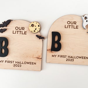 May include: Two wooden signs with the text "Our Little" and the letter "B" in black. The signs also have the text "My First Halloween 2023" and Halloween-themed decorations such as bats, a moon, and ghosts.