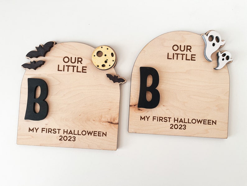 May include: Two wooden signs with the text "Our Little" and the letter "B" in black. The signs also have the text "My First Halloween 2023" and Halloween-themed decorations such as bats, a moon, and ghosts.