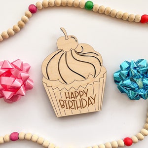 May include: Wooden cupcake cutout with the text "Happy Birthday" on it. The cutout is surrounded by a wooden bead garland with red, green, and yellow beads. There are two pink and blue bows on either side of the cutout.