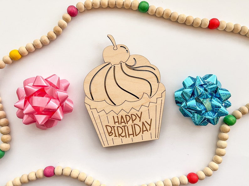 May include: Wooden cupcake cutout with the text "Happy Birthday" on it. The cutout is surrounded by a wooden bead garland with red, green, and yellow beads. There are two pink and blue bows on either side of the cutout.