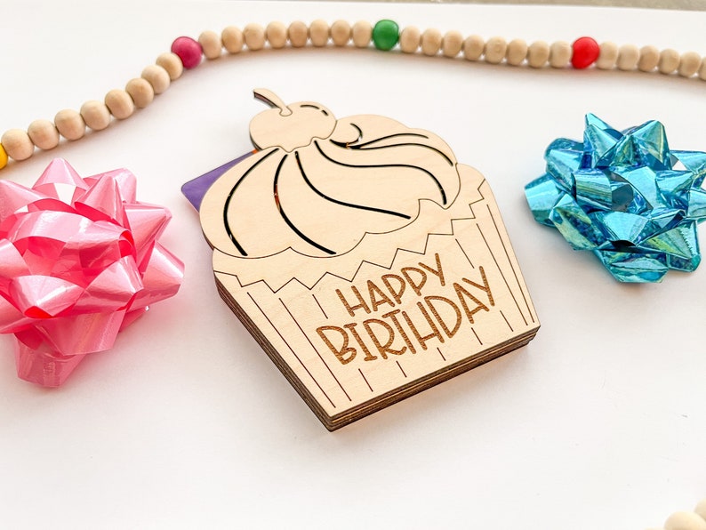 May include: A wooden cupcake card with the text "Happy Birthday" on it. The cupcake is decorated with a cherry on top and has a purple and white gift card inside.