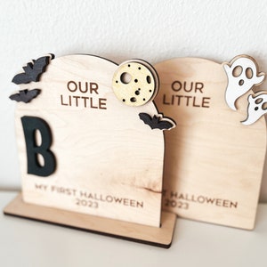 May include: Two wooden Halloween decorations with the text "Our Little" and a letter "B". The decorations are decorated with bats, ghosts, and a moon. The text "My First Halloween 2023" is on the bottom of the left decoration. The text "Halloween 2023" is on the bottom of the right decoration.