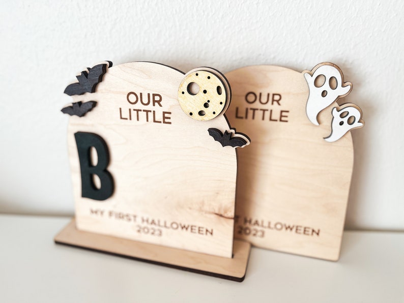 May include: Two wooden Halloween decorations with the text "Our Little" and a letter "B". The decorations are decorated with bats, ghosts, and a moon. The text "My First Halloween 2023" is on the bottom of the left decoration. The text "Halloween 2023" is on the bottom of the right decoration.