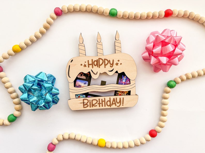 以下が含まれることがあります： 「Happy Birthday!」と書かれた木製のケーキ型のフォトホルダー。3本のろうそくと写真を入れるスロットがあります。ホルダーは、赤、緑、黄色のアクセントが付いた木製のビーズの紐で囲まれています。