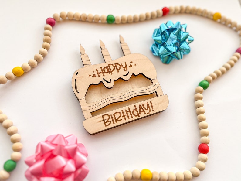 以下が含まれることがあります： ケーキの形をした木製バースデーカードで、「Happy Birthday!」の文字が書かれています。カードは、カラフルなアクセントが付いた木製のビーズの紐で囲まれています。