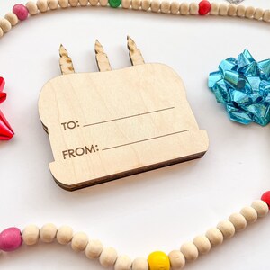 以下が含まれることがあります： 「To:」と「From:」の文字が書かれた木製の誕生日ケーキのギフトタグ。タグは、カラフルなアクセントが付いた木製のビーズの紐で囲まれています。