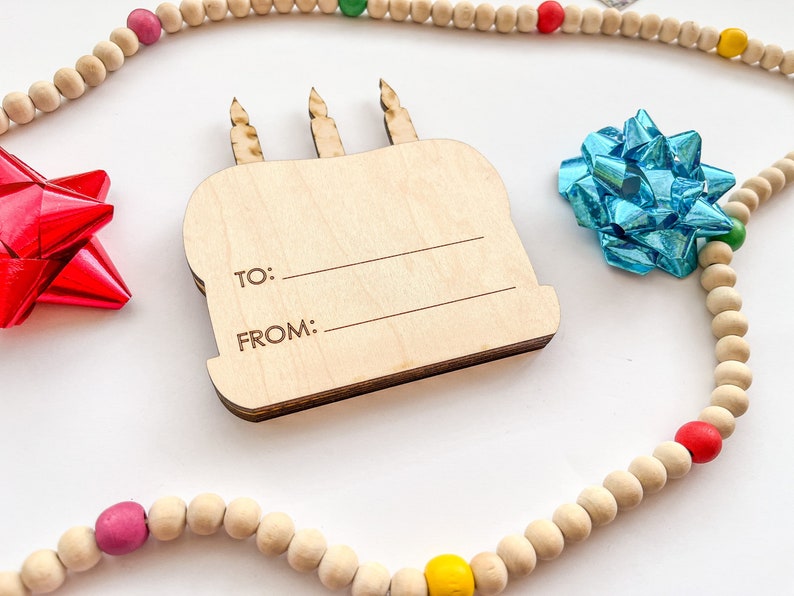 以下が含まれることがあります： 「To:」と「From:」の文字が書かれた木製の誕生日ケーキのギフトタグ。タグは、カラフルなアクセントが付いた木製のビーズの紐で囲まれています。