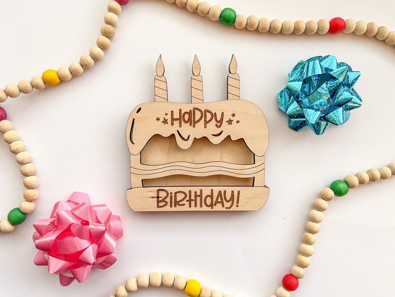 以下が含まれることがあります： ハッピーバースデー！と書かれた3本のろうそくがついた木製の誕生日ケーキの看板。看板はカラフルなビーズが置かれた白い背景の上に置かれています。