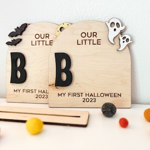 May include: Two wooden signs with the text "Our Little B" and "My First Halloween 2023". The signs are decorated with bats, ghosts, and a gold star.
