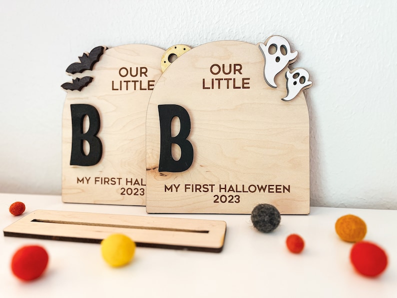 May include: Two wooden signs with the text "Our Little B" and "My First Halloween 2023". The signs are decorated with bats, ghosts, and a gold star.