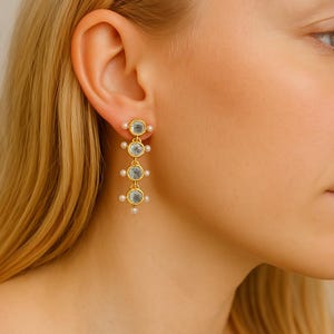 May include: Gold dangle earrings with four clear round stones and small white pearls. The earrings are hanging from a woman's ear.