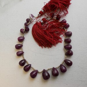 Könnte beinhalten: Eine Kette aus tiefvioletten Edelsteinperlen in Tropfenform, auf einer silbernen Schnur mit roten Quastenakzenten aufgefädelt.