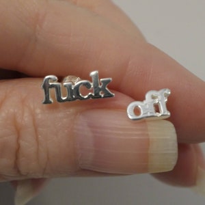 Può includere: Orecchini a bottone in argento con la scritta "fuck off". Gli orecchini sono in un carattere script e sono tenuti tra le dita. Gli orecchini sono piccoli e lucenti.