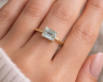 May include: A gold ring featuring a rectangular, light-green gemstone. The ring's band is a thin, polished gold. The gemstone is set with four prongs, and the ring is shown on a person's finger.