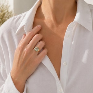 May include: A gold ring with a large, square-cut, light green gemstone. The ring is set in a simple gold band. The person is wearing a white button-down shirt.
