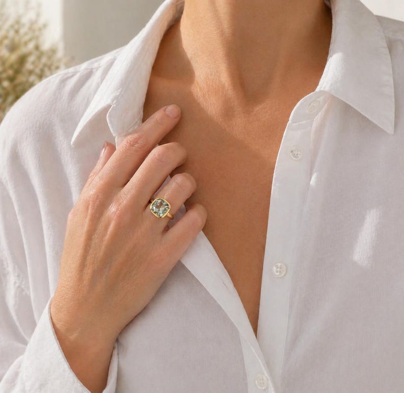 May include: A gold ring with a large, square-cut, light green gemstone. The ring is set in a simple gold band. The person is wearing a white button-down shirt.