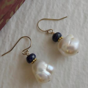 Könnte beinhalten: Ein Paar goldfarbene Ohrringe mit großen, unregelmäßigen weißen Perlen. Jeder Ohrring hat eine kleine, dunkelblaue Perle und einen goldfarbenen Akzent in der Nähe des Hakens.