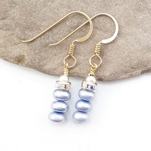 May include: A pair of dangle earrings featuring light blue, pearlescent beads. Each earring has three stacked beads, a silver-colored accent with clear crystals, and gold-colored ear wires. The earrings are displayed on a light gray stone surface.
