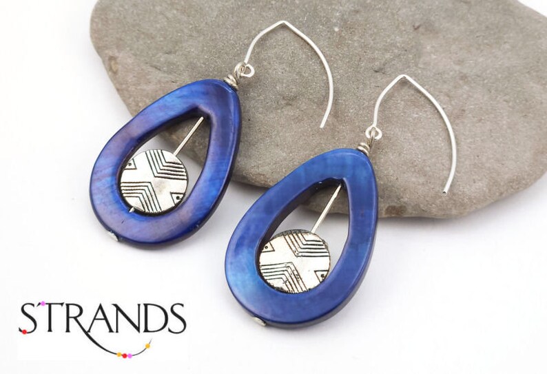 May include: A pair of blue teardrop earrings with a silver geometric design in the center. The earrings have a silver hook and are displayed on a gray stone. The word "STRANDS" is visible.