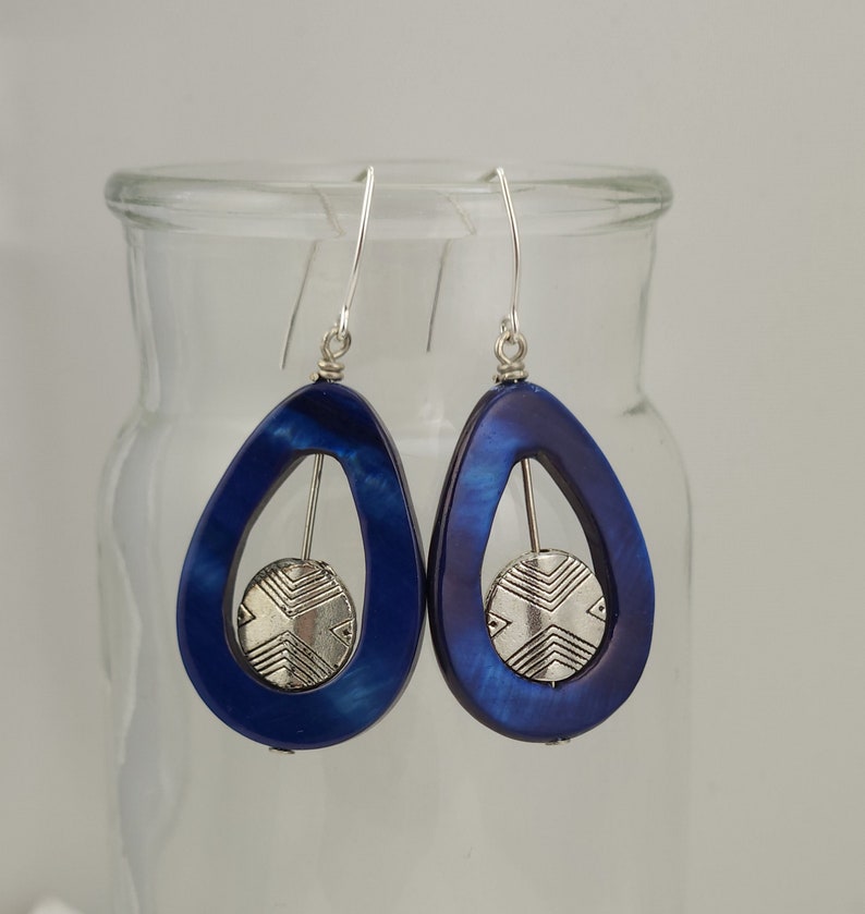 May include: A pair of teardrop-shaped earrings with a deep blue, iridescent finish. Each earring features a silver-colored, circular accent with a geometric design suspended within the teardrop frame. The earrings hang from silver-colored ear wires.