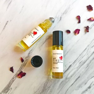 May include: Two glass bottles of natural perfume oil with rollerball applicators. The bottles are labelled "heartscent" and "MY FIRST natural perfume oil LAVENDER" with a red heart graphic. The bottles are on a white marble surface with dried rose petals.