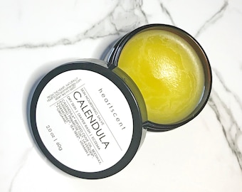 May include: A small, round jar of calendula skin nourishing salve, labeled "heartscent" and "Calendula" with a list of ingredients. The jar is open and the salve is a bright yellow color.
