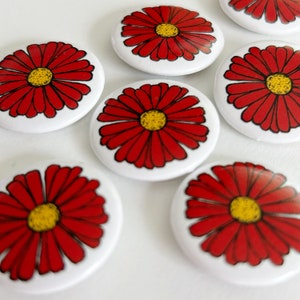 May include: White, round buttons with a red daisy design and yellow centers. The buttons are arranged on a white surface. The daisy petals are outlined in black.