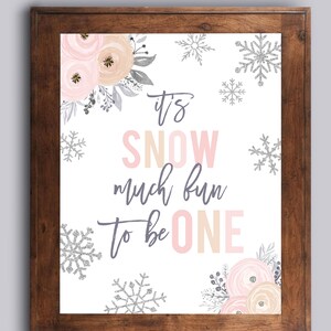 May include: A framed print with the text "It's SNOW much fun to be ONE" in pink, gray, and beige. The print is decorated with floral and snowflake designs in pink, silver, and gray. The frame is a dark brown color.