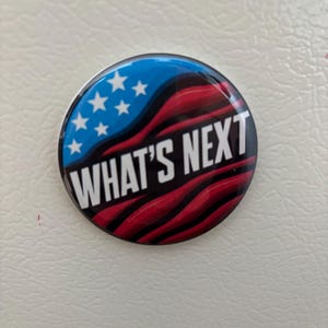 May include: A round button with a patriotic design. The top section is blue with white stars, transitioning to a red and black wavy pattern. The words "WHAT'S NEXT" are in white.