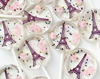 May include: A collection of round, clear lollipops on white sticks, each featuring a dark purple graphic of the Eiffel Tower, pink floral accents, and small dark blue sprinkles embedded within the hard candy surface.
